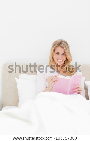 A closer shot showing a woman in bed sitting upright with a book in hand as she looks ahead and smiles.