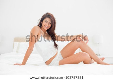 A woman who is smiling and sitting on the width of the bed with a raised knee and one arm placed across the knee while the other arm supports her.