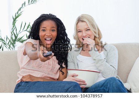 Two women are sitting down looking at the camera with popcorn and a television remote