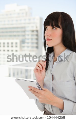 A pondering woman holds a notepad and puts a pen to her chin while in her office
