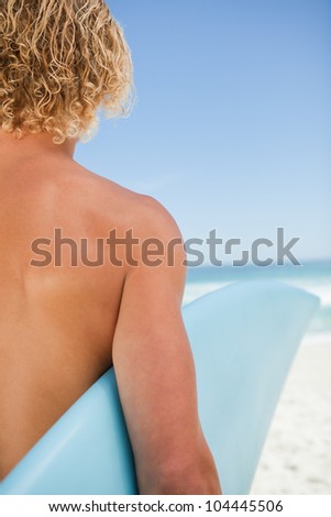 Rear view of a young blonde man standing on the beach while holding his surfboard