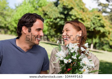 Man smiling as he watches his friend laughing while holding a bunch of flowers while sitting in a bright park