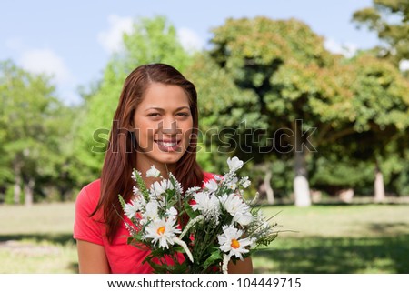 Woman standing in a bright park while looking into the camera and holding a bunch of flowers