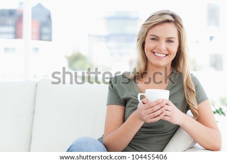 A woman sitting on the couch with a cup in her hands and smiling as she looks in front of her.