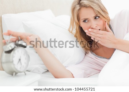 A woman in bed is leaning up and reaching to turn off the alarm as she yawns and checks the time