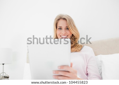 A low angle shot of a woman in bed with a tablet pc in hand as she is looking forward and smiling.