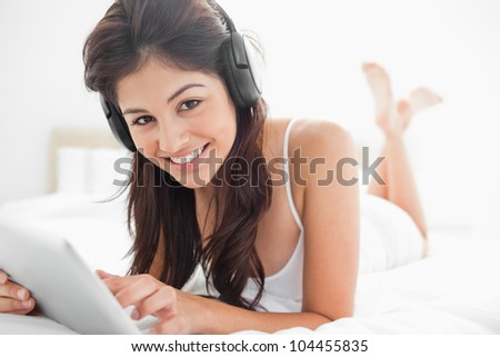 A smiling woman with her legs crossed, using both a tablet and headphones while looking forward.