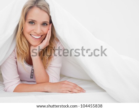 A woman at the end of the bed under the quilt, which is raised to her head level, as her hand supports her head.