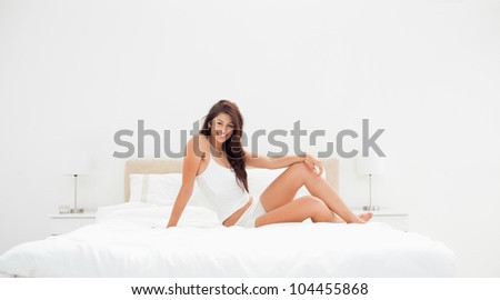 A wide angle shot, showing a woman sitting across the width of the bed, with a knee raised, smiling. it also shows the entire room.