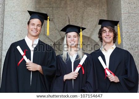 Graduates holding their diploma while posing with university in background