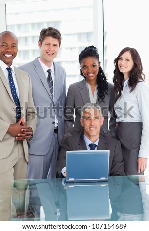 Smiling manager sitting at the desk behind a laptop and surrounded by his team
