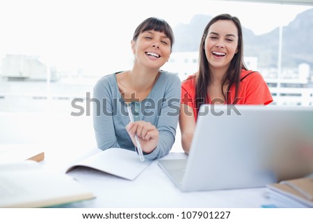 A girl and her friend both sitting together in front of the laptop laughing as they look into the camera