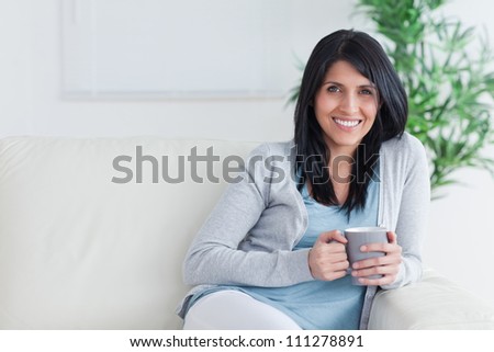 Smiling woman holding a mug with two hands while resting on a sofa in a living room