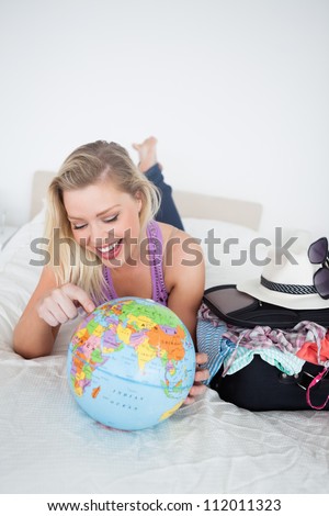 Student with a suitcase pointing on a globe while lying on her bed