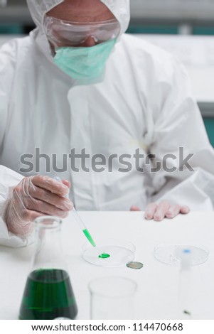 Chemist in protective suit adding green liquid to petri dish in the lab