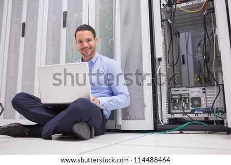 Smiling man sitting on floor checking servers with laptop in data center