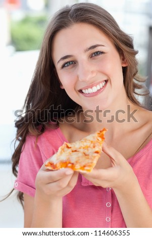 A close up shot of a woman looking into the camera as she holds a piece of pizza in her hand