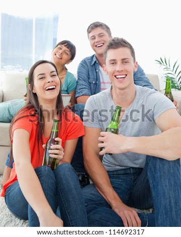 A group of friends sitting on the couch and the floor as they hold beers in their hands while laughing