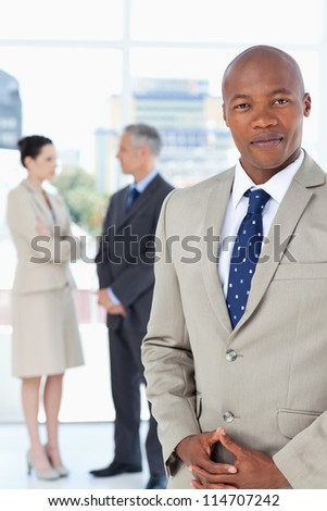 Young executive wearing a suit and crossing his hands with his team behind him