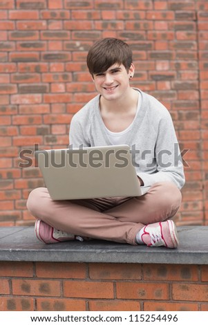 Young man sitting cross legged on wall with laptop in college
