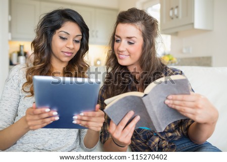 Two girls reading a book and holding a tablet computer while sitting in a couch