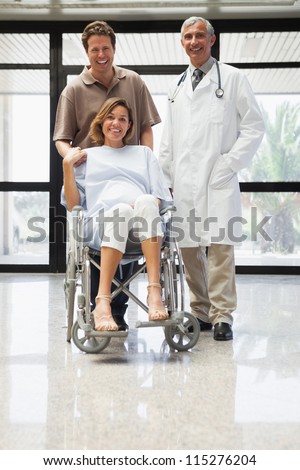 Pregnant woman in wheelchair, partner and doctor smiling in hospital corridor