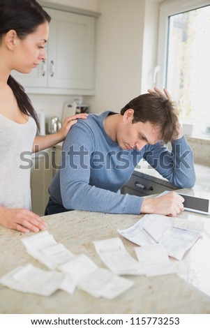 Wife comforting stressed husband over bills in kitchen with tablet pc