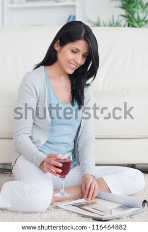 Smiling woman turning pages of a magazine while holding a glass of red wine in a living room