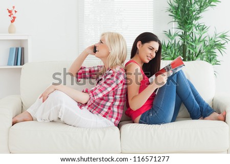 Women sitting back to back on couch, one is looking at magazine, one is on mobile phone