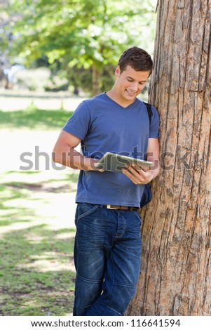 male student leaning against a tree while using a touch pad in a park