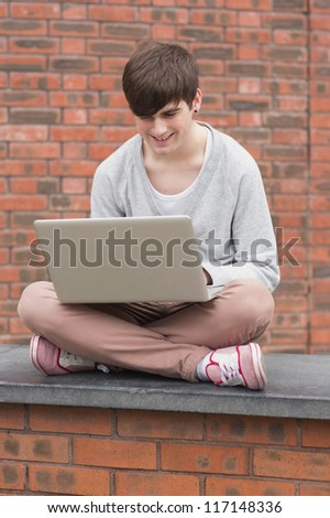 Man sitting cross legged on wall and using laptop outside in college