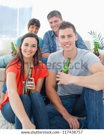 A smiling group of friends as one couple sit on the floor and the other couple sit on the couch with beers