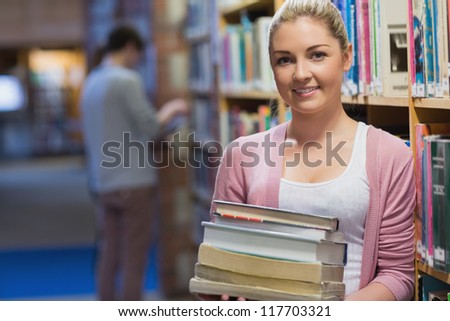 Woman holding pile of books in college library beside bookshelf