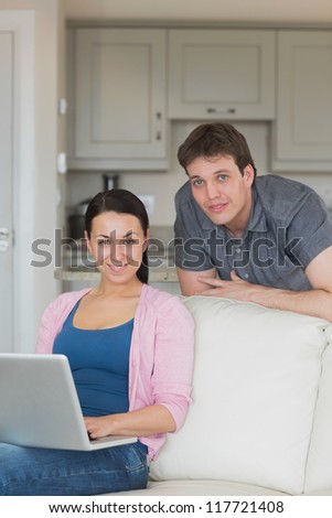 Woman sitting on the couch and uses her laptop while the man stands behind her