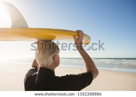 Rear view of senior man carrying surfboard on head at beach during sunny day