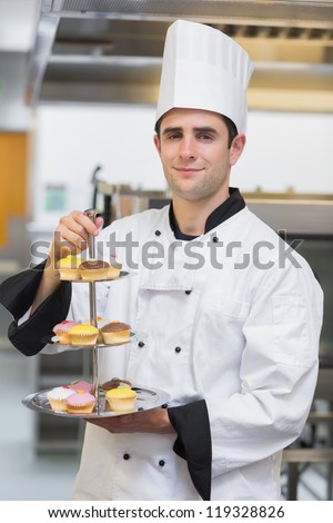 Baker smiling and holding tiered cake tray with cupcakes in kitchen