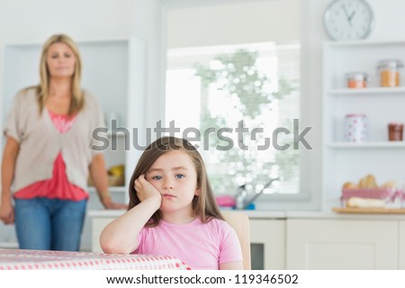 Child sitting at kitchen table looking angry with mother watching on