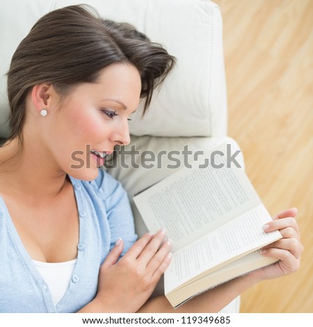 Smiling brunette reading a novel lying on the sofa in the living room