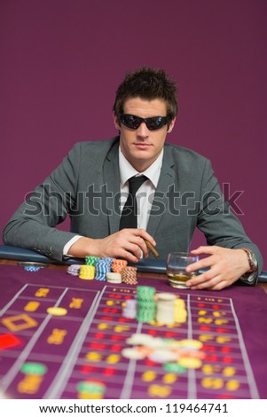 Man wearing sun glasses at roulette table drinking whiskey and smoking cigar