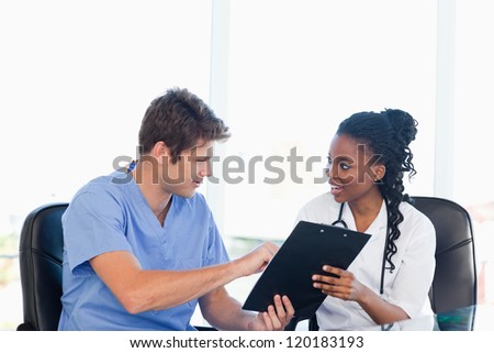 Young serious doctor showing something on a clipboard to his smiling co-worker