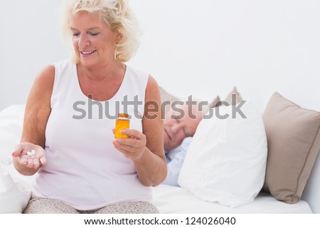Aged woman with the opened pill bottle while a man sleeping on the bed