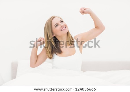 Sleepy smiling young woman waking up in bed and stretching her arms up