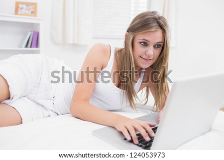 Blonde woman typing on the laptop lying on her bed  in the white bedroom