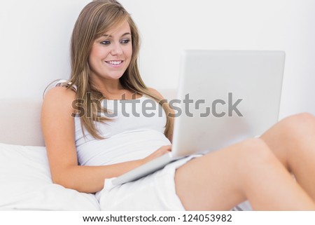 Blond woman sitting on the bed with her laptop in the white bedroom