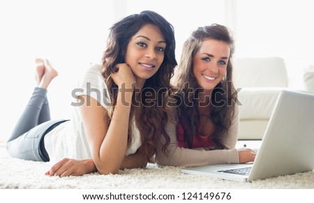 Two smiling women typing on a computer while lying on the floor in a living room