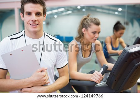 Male spinning class instructor holding clipboard with two women on exercise bikes