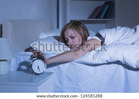 Irritated young woman in bed extending hand to alarm clock at home