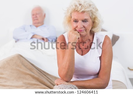 Discouraged old woman sitting on the bed with old man sleeping