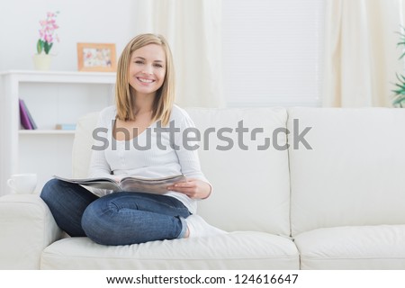 Portrait of happy young woman with magazine sitting on couch at home