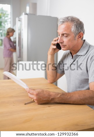 Mature man calling with a sheet of paper while his wife cooking in the kitchen